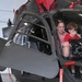 A woman and a young child are seated inside the cockpit of a military helicopter, likely during an open house or public display event at an aviation facility. The helicopter appears to be a dark gray/black military rotorcraft, possibly an AH-6 or similar light attack helicopter, parked inside a hangar. The child and adult appear to be exploring the cockpit instrumentation.