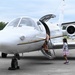 A white private/military jet is parked on a tarmac with its door open and stairs deployed. A group of people including children, a civilian adult, and a uniformed military service member are gathered near the aircraft's entrance. The setting appears to be a small airfield or military airbase, likely during an open house or familiarization event.