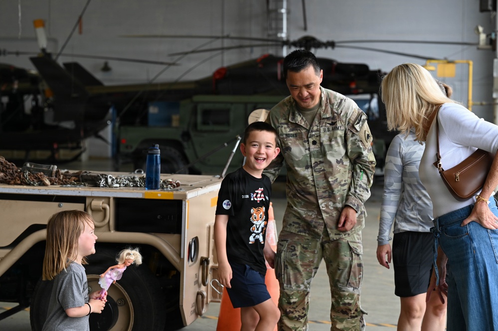 A U.S. Army soldier in MultiCam/OCP camouflage uniform, identified by name tape as 'ROOKS,' interacts joyfully with children and a woman in what appears to be a military hangar. The scene suggests a family reunion or homecoming event, with a laughing boy and a young girl holding a doll visible in the foreground. Military vehicles including a Humvee and helicopters are visible in the background.