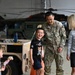A U.S. Army soldier in MultiCam/OCP camouflage uniform, identified by name tape as 'ROOKS,' interacts joyfully with children and a woman in what appears to be a military hangar. The scene suggests a family reunion or homecoming event, with a laughing boy and a young girl holding a doll visible in the foreground. Military vehicles including a Humvee and helicopters are visible in the background.