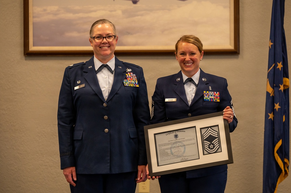 Two U.S. Air Force personnel in dress blue uniforms pose together during a promotion ceremony. The individual on the right, identified by name tag as 'Keiser,' holds a framed Certificate of Promotion alongside a Chief Master Sergeant rank insignia display. The ceremony appears to be an official military promotion event.