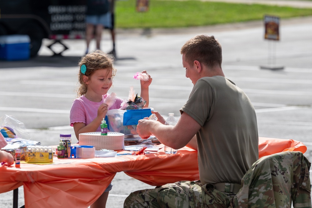 A young girl in a pink shirt and a person in military camouflage uniform are seated together at an outdoor craft table covered with an orange tablecloth. They appear to be engaged in a craft activity involving feathers, glue, and various art supplies. The setting appears to be an outdoor community or military family event.