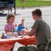 A young girl in a pink shirt and a person in military camouflage uniform are seated together at an outdoor craft table covered with an orange tablecloth. They appear to be engaged in a craft activity involving feathers, glue, and various art supplies. The setting appears to be an outdoor community or military family event.