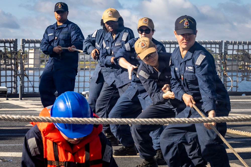 A group of U.S. Navy sailors in dark blue coveralls are pulling a thick mooring rope on the deck of a naval vessel. The caps worn by several sailors display 'USS St. Louis LCS 19' insignia, identifying the ship. One sailor in the foreground wears a blue hard hat and orange life vest, likely indicating a safety or supervisory role during the line-handling evolution.