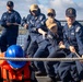 A group of U.S. Navy sailors in dark blue coveralls are pulling a thick mooring rope on the deck of a naval vessel. The caps worn by several sailors display 'USS St. Louis LCS 19' insignia, identifying the ship. One sailor in the foreground wears a blue hard hat and orange life vest, likely indicating a safety or supervisory role during the line-handling evolution.