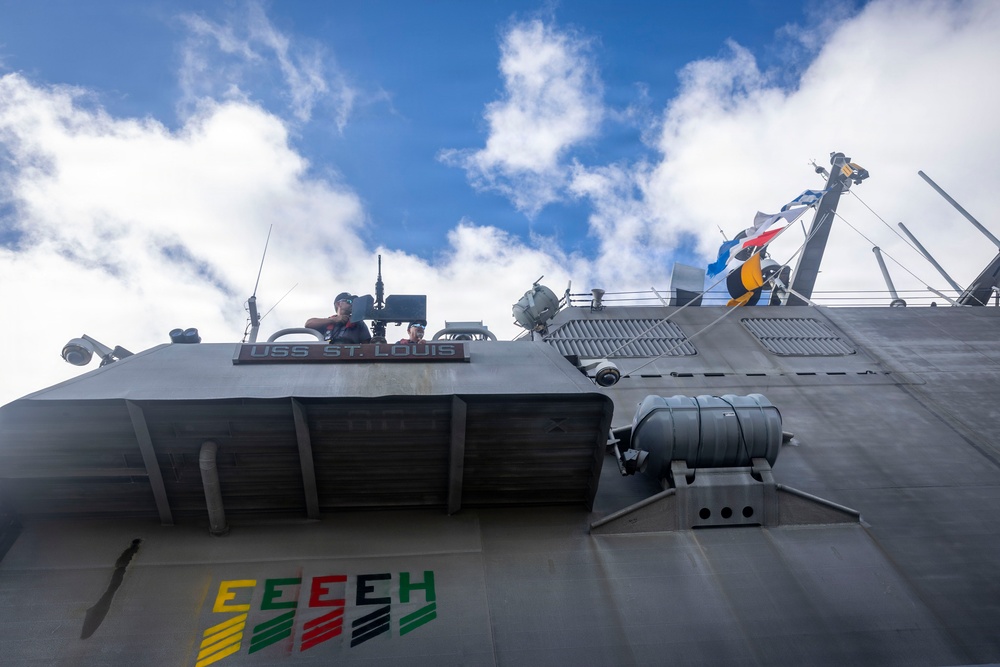 A low-angle upward shot of the USS St. Louis, a U.S. Navy vessel, showing its superstructure against a partly cloudy blue sky. A crew member is visible at an elevated station near the bridge, and naval signal flags are flying from a mast. The hull displays colorful efficiency award markings near the waterline area.