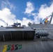 A low-angle upward shot of the USS St. Louis, a U.S. Navy vessel, showing its superstructure against a partly cloudy blue sky. A crew member is visible at an elevated station near the bridge, and naval signal flags are flying from a mast. The hull displays colorful efficiency award markings near the waterline area.