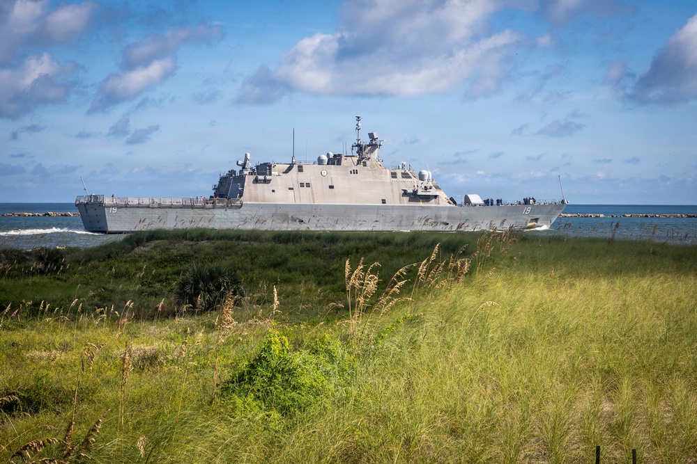 A U.S. Navy Littoral Combat Ship bearing hull number 19 is transiting a coastal waterway, photographed from a shoreline with sea grass and coastal vegetation in the foreground. The vessel appears to be underway in calm conditions with personnel visible on deck. The image was taken from a low vantage point near a beach or inlet, giving a dramatic perspective of the warship against a partly cloudy sky.