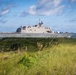 A U.S. Navy Littoral Combat Ship bearing hull number 19 is transiting a coastal waterway, photographed from a shoreline with sea grass and coastal vegetation in the foreground. The vessel appears to be underway in calm conditions with personnel visible on deck. The image was taken from a low vantage point near a beach or inlet, giving a dramatic perspective of the warship against a partly cloudy sky.