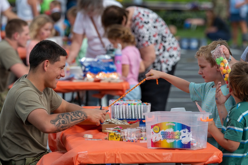 A community arts and crafts event is taking place outdoors at tables covered with orange tablecloths. A young man with a tattoo on his arm, wearing an olive green t-shirt, is smiling and engaging with two children in a craft activity. The table is stocked with markers, beads, and an 'Arts &amp; Crafts Collection' kit.