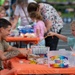 A community arts and crafts event is taking place outdoors at tables covered with orange tablecloths. A young man with a tattoo on his arm, wearing an olive green t-shirt, is smiling and engaging with two children in a craft activity. The table is stocked with markers, beads, and an 'Arts &amp; Crafts Collection' kit.