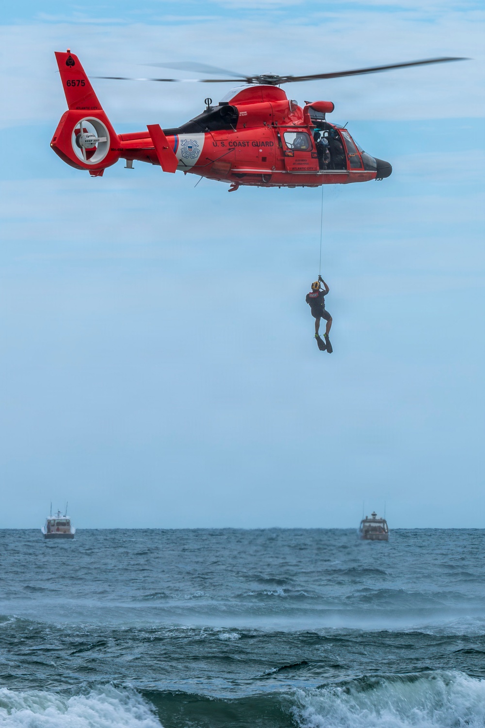 A U.S. Coast Guard helicopter (tail number 6575, based in Atlantic City) hovers over open ocean water while a rescue swimmer is being lowered or hoisted on a cable. Two small vessels are visible on the horizon in the background. This appears to be either a rescue operation or a training exercise.