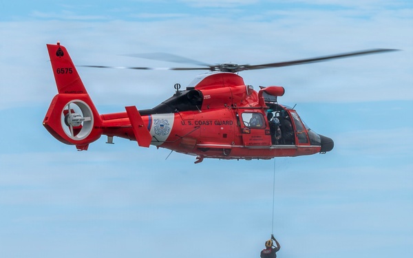 A U.S. Coast Guard helicopter (tail number 6575, based in Atlantic City) hovers over open ocean water while a rescue swimmer is being lowered or hoisted on a cable. Two small vessels are visible on the horizon in the background. This appears to be either a rescue operation or a training exercise.