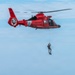A U.S. Coast Guard helicopter (tail number 6575, based in Atlantic City) hovers over open ocean water while a rescue swimmer is being lowered or hoisted on a cable. Two small vessels are visible on the horizon in the background. This appears to be either a rescue operation or a training exercise.