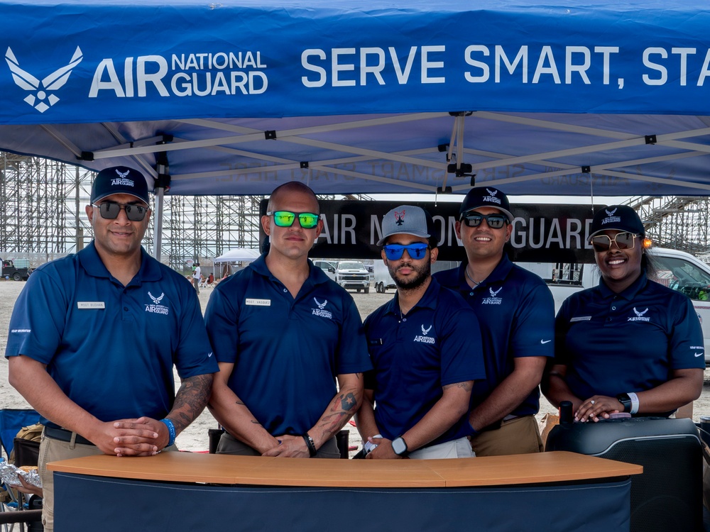Five Air National Guard recruiters or representatives are posing together behind a table at an outdoor recruitment booth. They are all wearing matching navy blue Air National Guard polo shirts and sunglasses, standing under a blue branded canopy tent. The setting appears to be an outdoor event, possibly an airshow, with large hangar structures visible in the background.