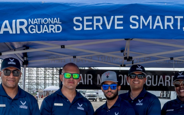 Five Air National Guard recruiters or representatives are posing together behind a table at an outdoor recruitment booth. They are all wearing matching navy blue Air National Guard polo shirts and sunglasses, standing under a blue branded canopy tent. The setting appears to be an outdoor event, possibly an airshow, with large hangar structures visible in the background.