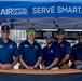 Five Air National Guard recruiters or representatives are posing together behind a table at an outdoor recruitment booth. They are all wearing matching navy blue Air National Guard polo shirts and sunglasses, standing under a blue branded canopy tent. The setting appears to be an outdoor event, possibly an airshow, with large hangar structures visible in the background.