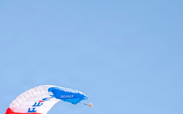 A skydiver is descending under a red, white, and blue parachute branded with 'RE/MAX' on the canopy. The jumper is trailing a long red, white, and blue striped banner or flag behind them against a clear blue sky. The scene appears to be a demonstration or airshow performance.