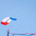 A skydiver is descending under a red, white, and blue parachute branded with 'RE/MAX' on the canopy. The jumper is trailing a long red, white, and blue striped banner or flag behind them against a clear blue sky. The scene appears to be a demonstration or airshow performance.