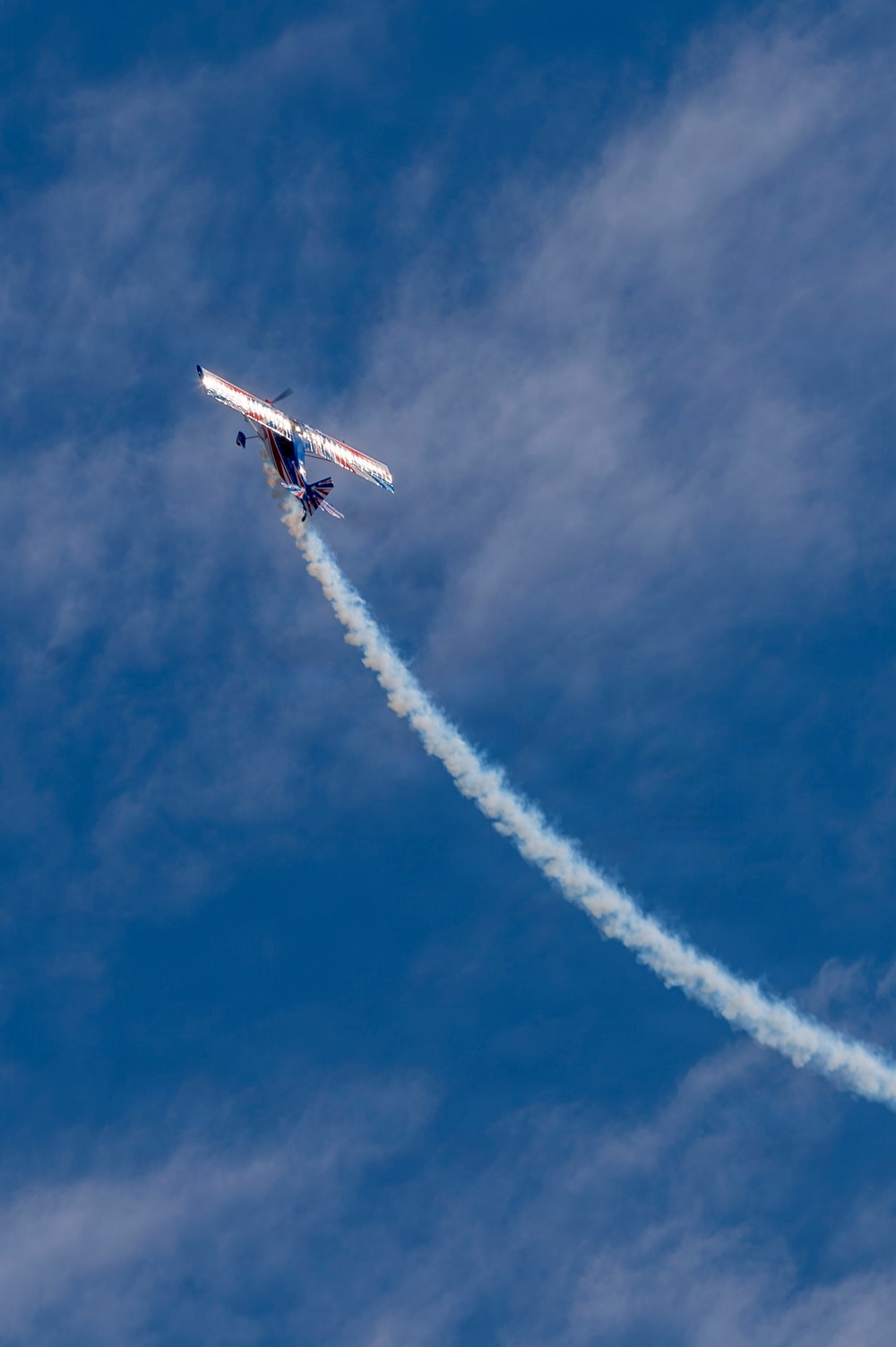A single-engine aerobatic propeller aircraft is captured mid-maneuver in a blue sky with scattered clouds. The aircraft is banking steeply and emitting a white smoke trail, consistent with an airshow performance. A second smoke trail is visible crossing the first, suggesting a prior pass or maneuver.