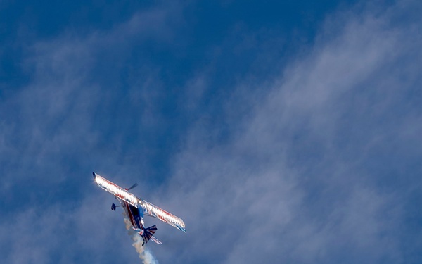 A single-engine aerobatic propeller aircraft is captured mid-maneuver in a blue sky with scattered clouds. The aircraft is banking steeply and emitting a white smoke trail, consistent with an airshow performance. A second smoke trail is visible crossing the first, suggesting a prior pass or maneuver.