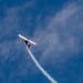 A single-engine aerobatic propeller aircraft is captured mid-maneuver in a blue sky with scattered clouds. The aircraft is banking steeply and emitting a white smoke trail, consistent with an airshow performance. A second smoke trail is visible crossing the first, suggesting a prior pass or maneuver.
