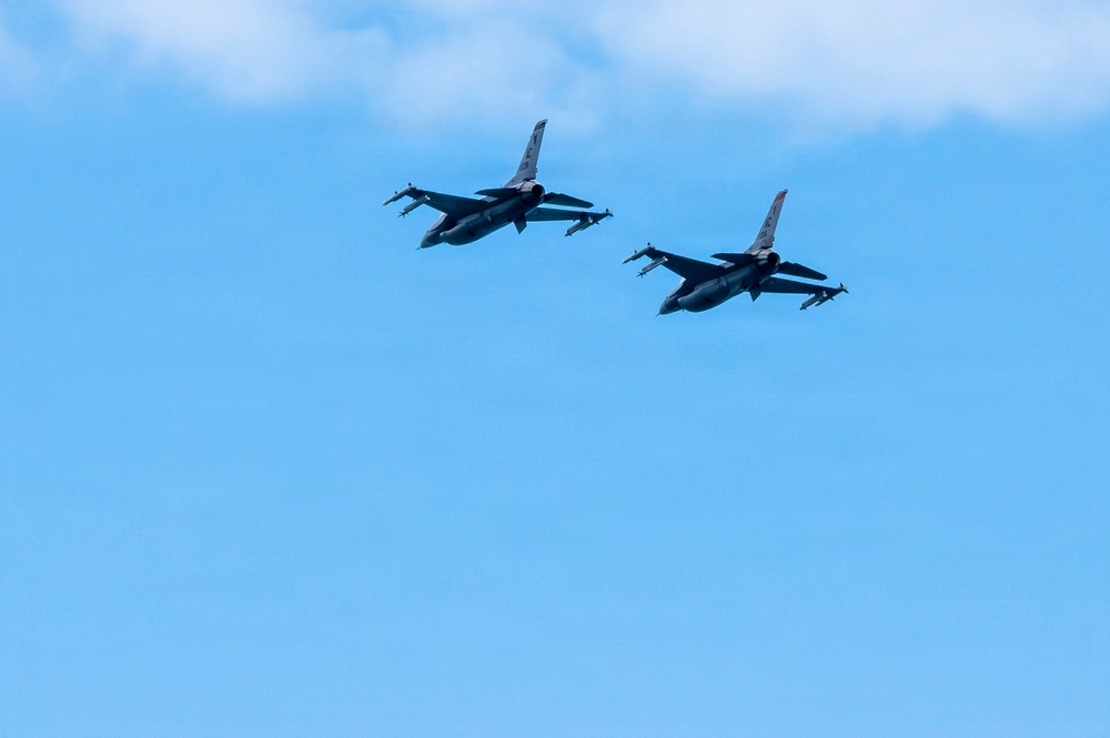 Two military fighter jets are flying in formation against a mostly clear blue sky with light clouds in the upper right corner. The aircraft appear to be F-16 Fighting Falcons based on their distinctive silhouette, flying in a loose echelon formation. Both aircraft appear to be carrying external stores such as missiles or fuel tanks under their wings.