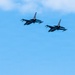 Two military fighter jets are flying in formation against a mostly clear blue sky with light clouds in the upper right corner. The aircraft appear to be F-16 Fighting Falcons based on their distinctive silhouette, flying in a loose echelon formation. Both aircraft appear to be carrying external stores such as missiles or fuel tanks under their wings.