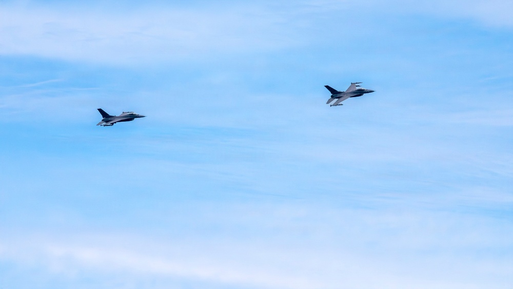 Two military fighter jets are flying in formation against a blue sky with light cirrus clouds. The aircraft appear to be F-16 Fighting Falcons based on their distinctive delta wing and tail configuration. They are flying in a loose trail or echelon formation at similar altitude.