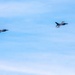 Two military fighter jets are flying in formation against a blue sky with light cirrus clouds. The aircraft appear to be F-16 Fighting Falcons based on their distinctive delta wing and tail configuration. They are flying in a loose trail or echelon formation at similar altitude.