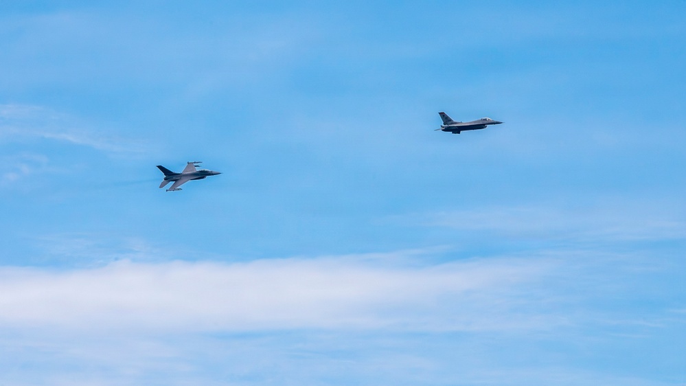 Two military fighter jets are flying in formation against a blue sky with light cloud cover. Both aircraft appear to be F-16 Fighting Falcons based on their distinctive delta wing and tail configuration. The jets are photographed at a distance, flying in a loose formation at similar altitudes.