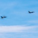 Two military fighter jets are flying in formation against a blue sky with light cloud cover. Both aircraft appear to be F-16 Fighting Falcons based on their distinctive delta wing and tail configuration. The jets are photographed at a distance, flying in a loose formation at similar altitudes.