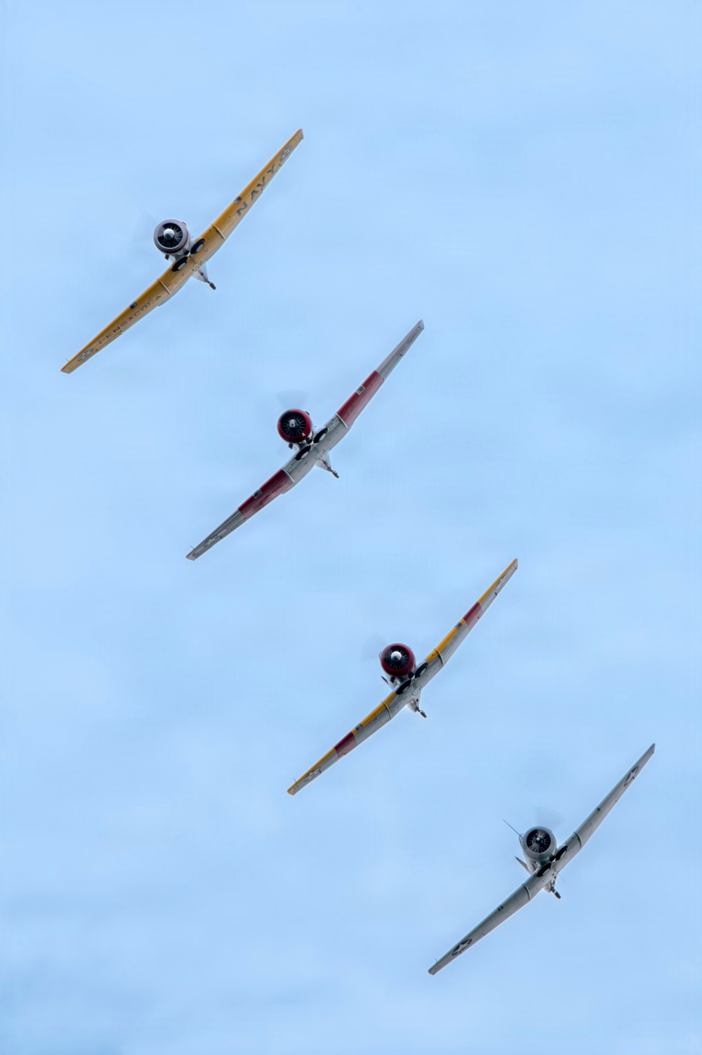Four vintage propeller-driven trainer aircraft are flying in a diagonal echelon formation against a pale blue sky. The aircraft appear to be North American T-6 Texan (SNJ) trainers in various color schemes including yellow, red-and-white, yellow-and-red, and silver/natural metal. The lead aircraft bears visible 'NAVY' markings, suggesting this is a warbird airshow formation flight.