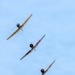 Four vintage propeller-driven trainer aircraft are flying in a diagonal echelon formation against a pale blue sky. The aircraft appear to be North American T-6 Texan (SNJ) trainers in various color schemes including yellow, red-and-white, yellow-and-red, and silver/natural metal. The lead aircraft bears visible 'NAVY' markings, suggesting this is a warbird airshow formation flight.