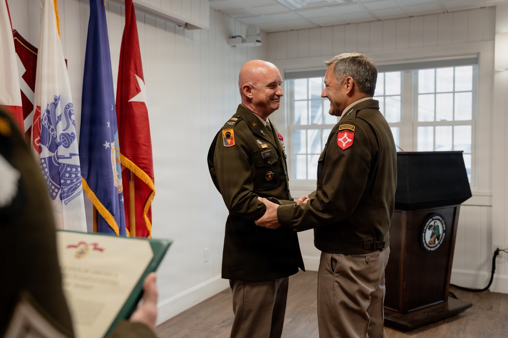 Two U.S. Army officers in olive green service uniforms are shaking hands and smiling during what appears to be a formal military ceremony or award presentation. A third person in the foreground holds a green folder containing what appears to be a certificate or citation. Multiple military flags and a podium with an official seal are visible in the background.