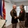 Col. Finch Concludes Distinguished Career, Leaves Lasting Legacy as Command State Chaplain Two U.S. Army officers in olive green service uniforms are shaking hands and smiling during what appears to be a formal military ceremony or award presentation. A third person in the foreground holds a green folder containing what appears to be a certificate or citation. Multiple military flags and a podium with an official seal are visible in the background.