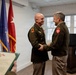 Two U.S. Army officers in olive green service uniforms are shaking hands and smiling during what appears to be a formal military ceremony or award presentation. A third person in the foreground holds a green folder containing what appears to be a certificate or citation. Multiple military flags and a podium with an official seal are visible in the background.