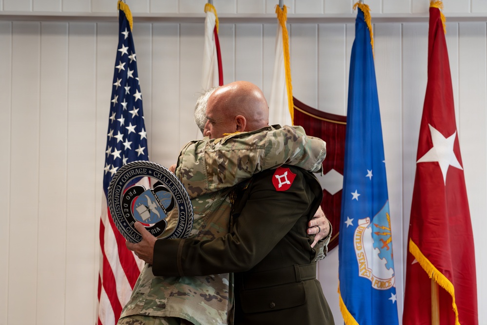 Two military personnel in uniform are embracing during what appears to be a formal military ceremony. One individual is wearing Army Combat Uniform (ACU/OCP) with a unit patch visible on the sleeve, while the other is in a dark military jacket. A commemorative medallion or plaque is visible being held during the embrace.