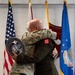Two military personnel in uniform are embracing during what appears to be a formal military ceremony. One individual is wearing Army Combat Uniform (ACU/OCP) with a unit patch visible on the sleeve, while the other is in a dark military jacket. A commemorative medallion or plaque is visible being held during the embrace.