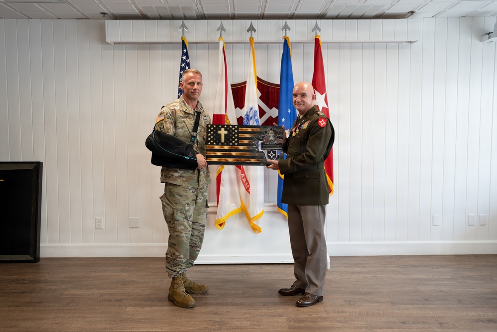Two military personnel are posing together while jointly holding a wooden plaque shaped like an American flag with a cross design. One individual is wearing U.S. Army combat uniform (OCP) with their arm in a sling, while the other is wearing a formal green dress uniform with medals and rank insignia. They are standing in front of multiple military flags in what appears to be an official ceremony or presentation setting.
