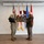 Col. Finch Concludes Distinguished Career, Leaves Lasting Legacy as Command State Chaplain Two military personnel are posing together while jointly holding a wooden plaque shaped like an American flag with a cross design. One individual is wearing U.S. Army combat uniform (OCP) with their arm in a sling, while the other is wearing a formal green dress uniform with medals and rank insignia. They are standing in front of multiple military flags in what appears to be an official ceremony or presentation setting.
