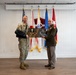Two military personnel are posing together while jointly holding a wooden plaque shaped like an American flag with a cross design. One individual is wearing U.S. Army combat uniform (OCP) with their arm in a sling, while the other is wearing a formal green dress uniform with medals and rank insignia. They are standing in front of multiple military flags in what appears to be an official ceremony or presentation setting.