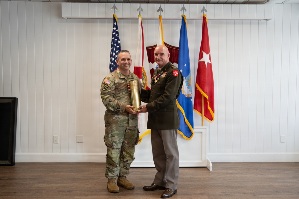 Two military personnel are posing together while jointly holding a large brass artillery shell casing, likely during a formal military ceremony or award presentation. One individual is wearing Army Combat Uniform (ACU/OCP pattern) and the other is in Army Service Uniform (green dress uniform) with multiple medals and insignia. Several flags are displayed in the background, including the U.S. flag, Florida state flag, Air Force flag, and a general officer flag with one star.