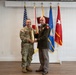 Two military personnel are posing together while jointly holding a large brass artillery shell casing, likely during a formal military ceremony or award presentation. One individual is wearing Army Combat Uniform (ACU/OCP pattern) and the other is in Army Service Uniform (green dress uniform) with multiple medals and insignia. Several flags are displayed in the background, including the U.S. flag, Florida state flag, Air Force flag, and a general officer flag with one star.