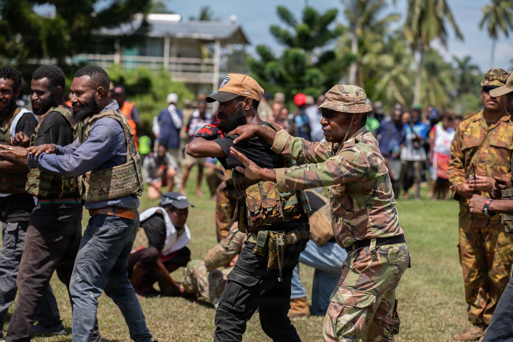 The image shows a tense scene involving multiple individuals in military/tactical camouflage uniforms engaged in what appears to be a physical confrontation or crowd control situation outdoors. Several people wearing camouflage gear and tactical vests are grappling with or restraining others on a grassy field. A crowd of onlookers is visible in the background near palm trees and a structure.
