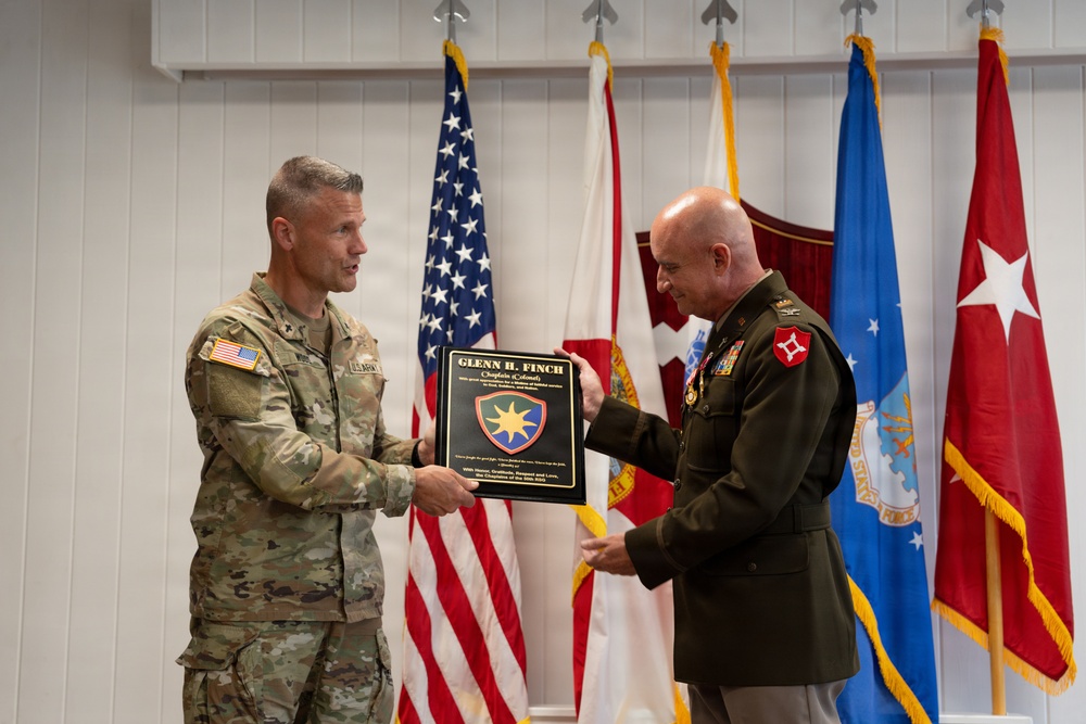 Two military officers are participating in a formal award or recognition ceremony, with one presenting a commemorative plaque to the other. The plaque bears the name 'Glenn H. Finch' and the title 'Chaplain (Colonel)'. The ceremony takes place in front of multiple military flags representing various branches of the U.S. Armed Forces.