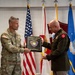 Two military officers are participating in a formal award or recognition ceremony, with one presenting a commemorative plaque to the other. The plaque bears the name 'Glenn H. Finch' and the title 'Chaplain (Colonel)'. The ceremony takes place in front of multiple military flags representing various branches of the U.S. Armed Forces.