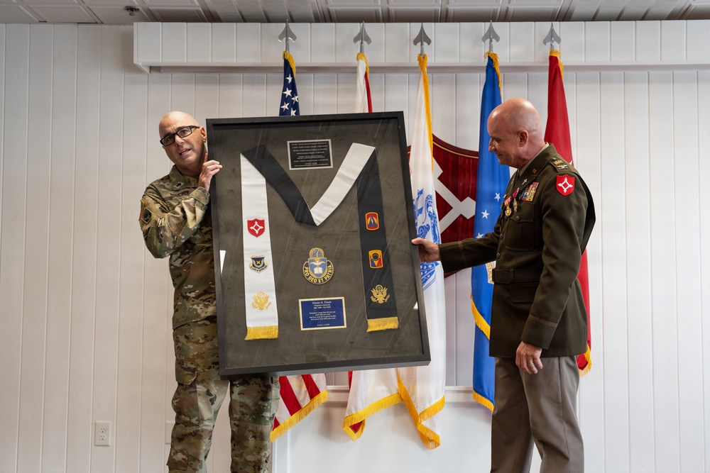 Two military personnel are holding up a framed shadow box display containing military insignia, patches, medals, and what appears to be a ceremonial stole or sash. The framed display includes multiple unit patches, a central medallion/crest, and a commemorative plaque, suggesting a retirement or award ceremony. Several military flags are visible in the background, including the U.S. flag and what appear to be Army unit flags.