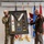 Col. Finch Concludes Distinguished Career, Leaves Lasting Legacy as Command State Chaplain Two military personnel are holding up a framed shadow box display containing military insignia, patches, medals, and what appears to be a ceremonial stole or sash. The framed display includes multiple unit patches, a central medallion/crest, and a commemorative plaque, suggesting a retirement or award ceremony. Several military flags are visible in the background, including the U.S. flag and what appear to be Army unit flags.