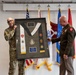 Two military personnel are holding up a framed shadow box display containing military insignia, patches, medals, and what appears to be a ceremonial stole or sash. The framed display includes multiple unit patches, a central medallion/crest, and a commemorative plaque, suggesting a retirement or award ceremony. Several military flags are visible in the background, including the U.S. flag and what appear to be Army unit flags.